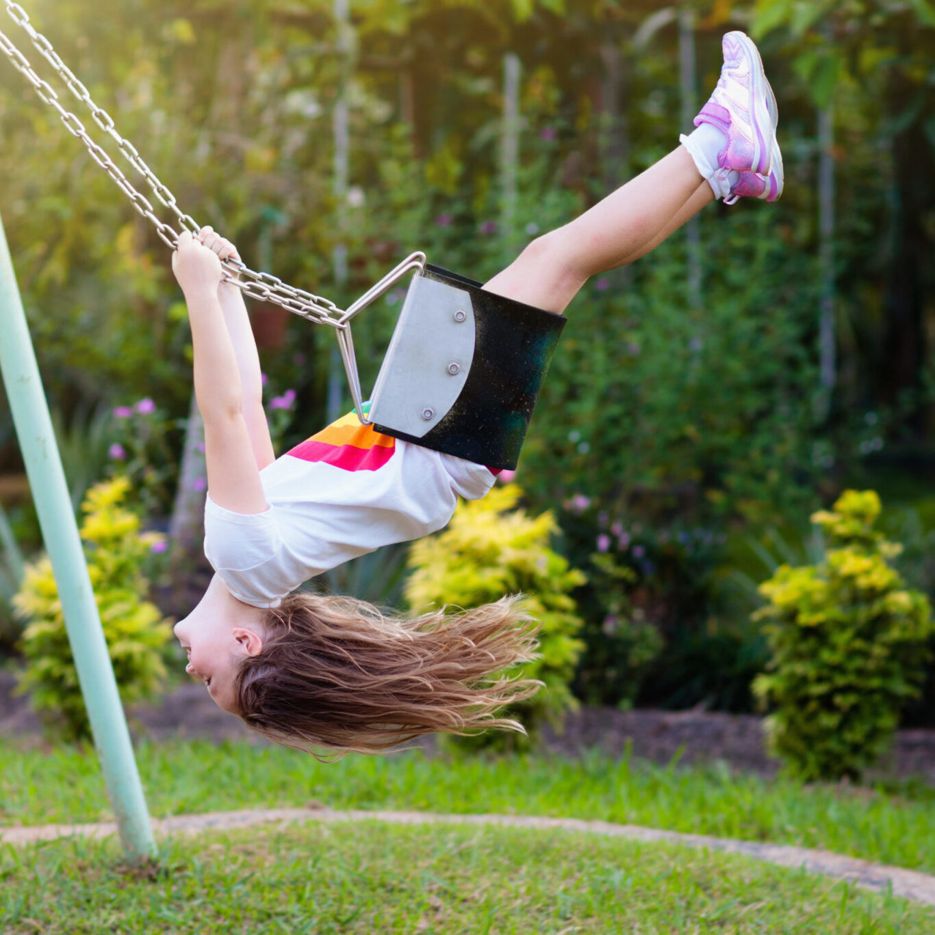 Child swinging on playground on sunny summer day in a park. Kids swing. School or kindergarten yard and play ground. Little girl flying high in the air on swing. Summer outdoor activity. Kid playing.